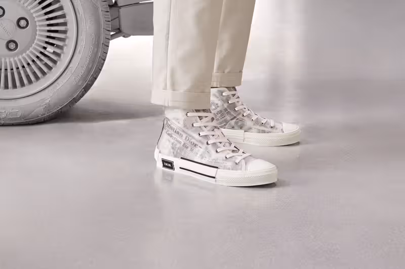 "Model wearing Dior B23 high-top sneakers with a black and white newspaper print; beige tailored trousers; translucent paneling and signature Dior rubber sole; luxury men's footwear editorial."