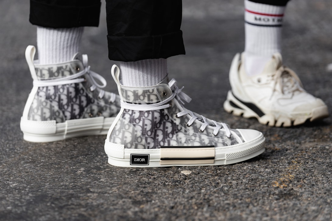 On-foot street style shot during fashion week featuring a person wearing Dior B23 high-top sneakers with a black and white Oblique monogram canvas upper under a clear translucent film; paired with cropped black trousers and white crew socks