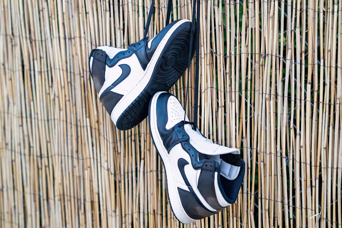 A pair of clean black and white Nike Air Jordan 1 high-top sneakers hanging by their long black laces to air dry outdoors. The shoes are suspended against a rustic, light brown vertical bamboo reed fence background.