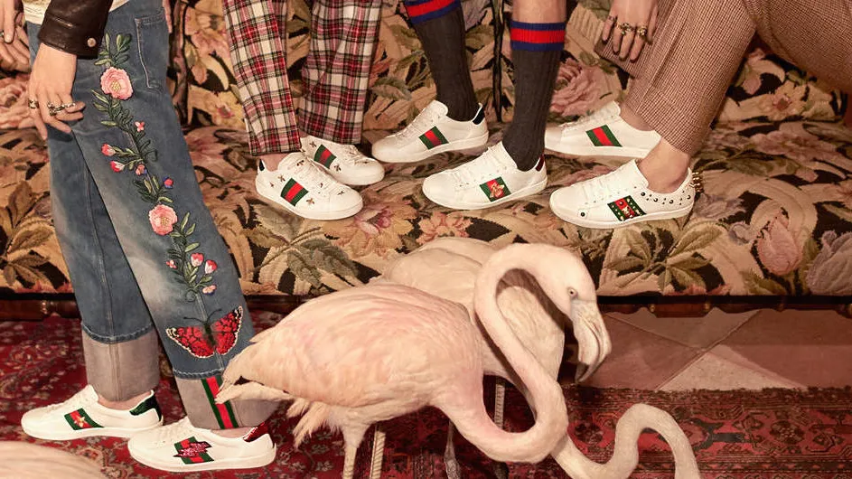 A pair of white leather Gucci Ace sneakers featuring the iconic metallic gold embroidered bee over the signature green and red web stripe, with metallic red and green leather heel tabs.