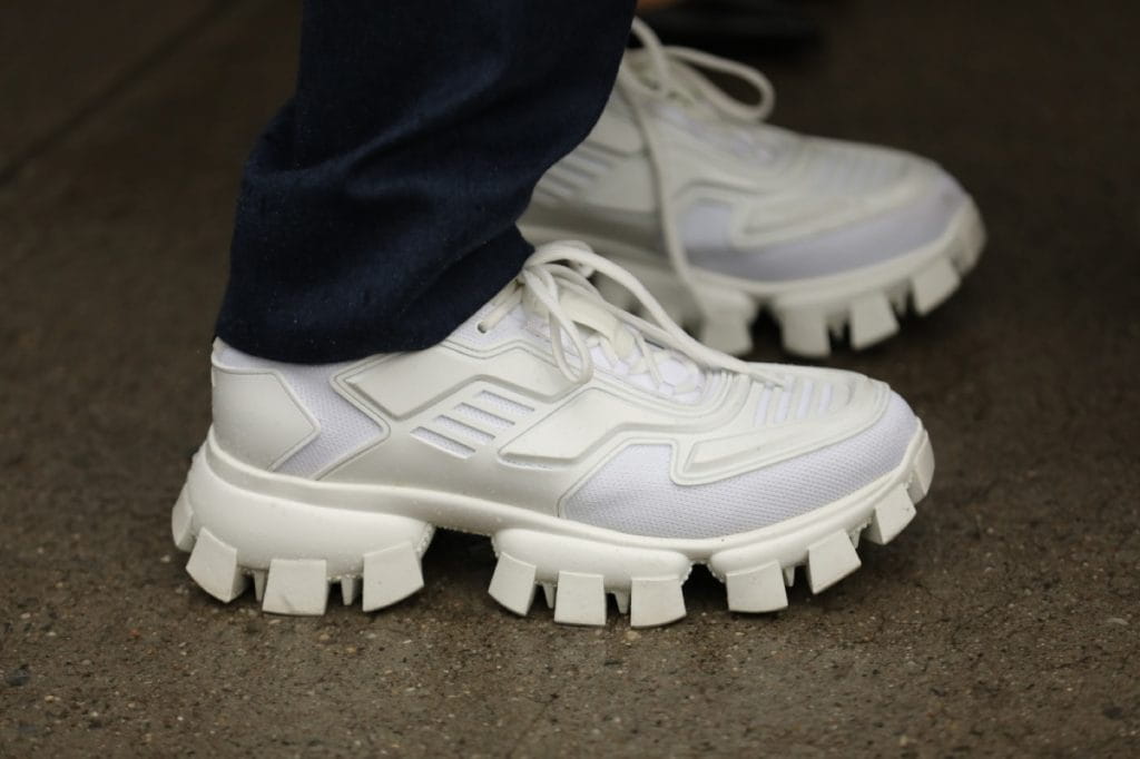 A close-up, low-angle shot of a person wearing a pair of all-white Prada Cloudbust Thunder sneakers paired with dark navy trousers. The sneakers feature an intricate, futuristic white mesh and rubber upper sitting atop a highly distinctive, oversized chunky sole with thick, blocky treading.