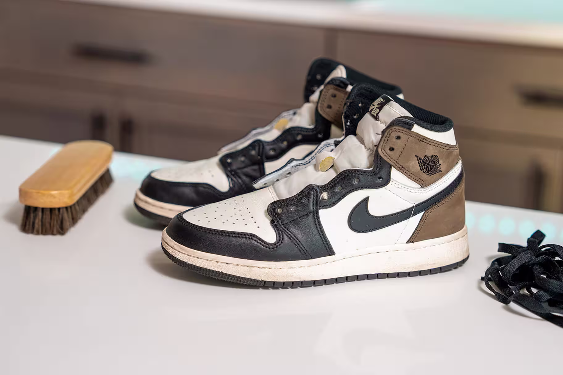A pair of unlaced black, white, and brown leather Nike Air Jordan 1 high-top sneakers resting on a white tabletop, prepared for cleaning.