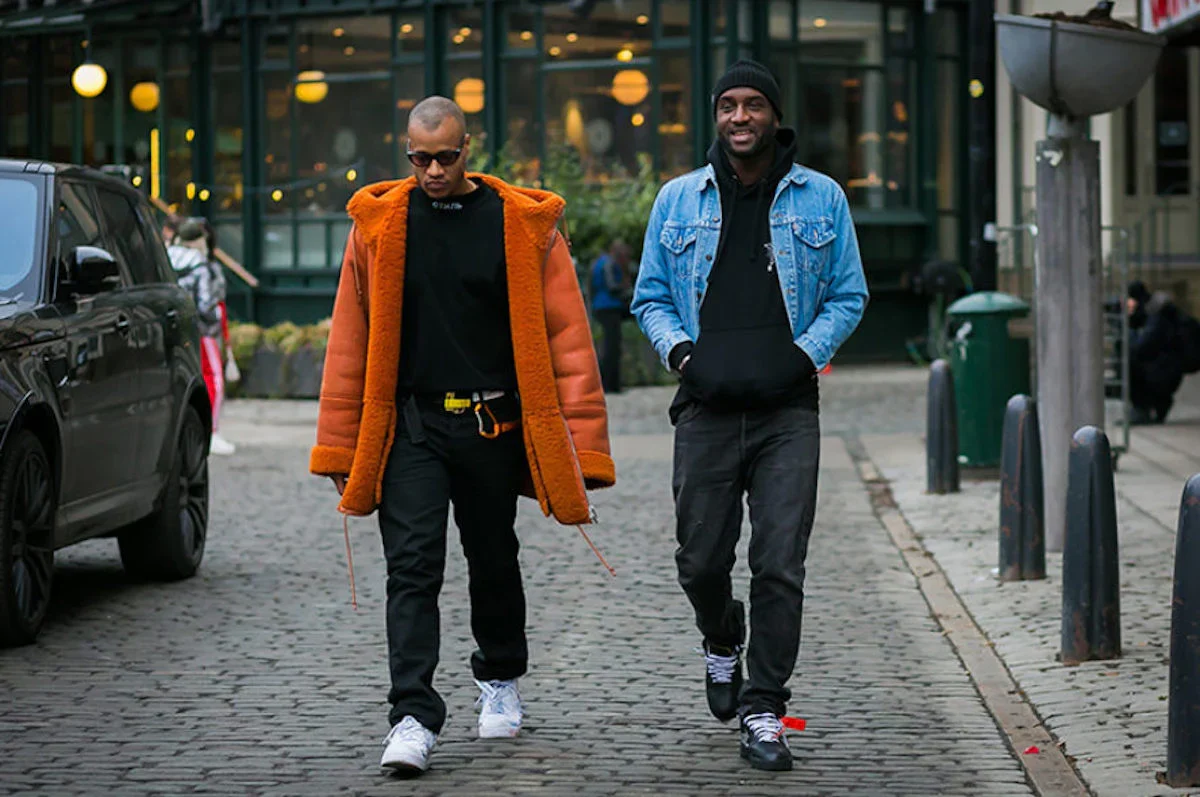 Designers Heron Preston and Virgil Abloh walking down a cobblestone street in urban winter streetwear; Heron Preston on the left wearing an oversized orange shearling coat over an all-black outfit, and Virgil Abloh on the right in a light wash denim jacket over a black hoodie and beanie with Off-White sneakers.